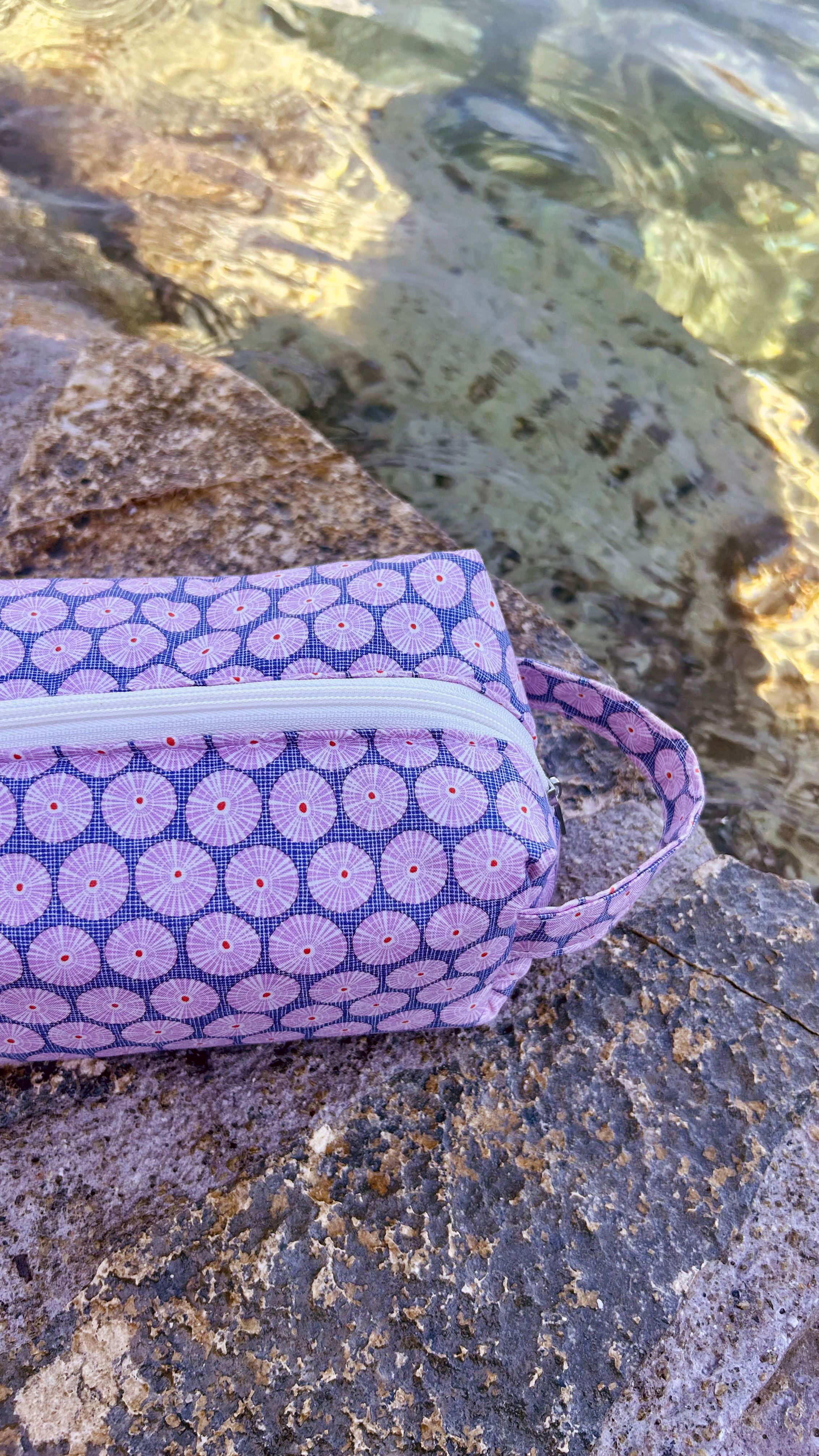 Lavender sea-urchin patterned cosmetic bag resting on a rock beside clear turquoise water