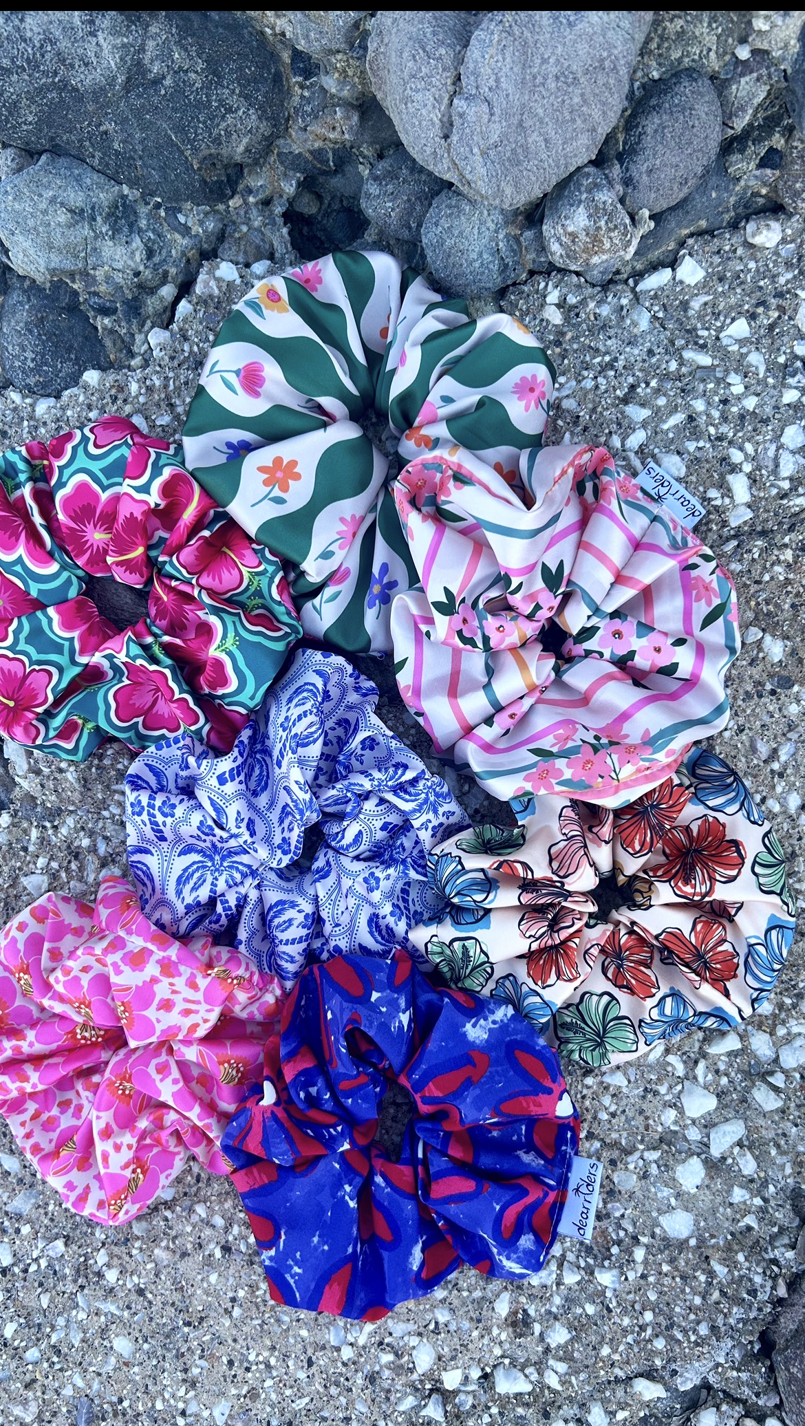 Colorful collection of floral and patterned scrunchies arranged on a pebbled beach