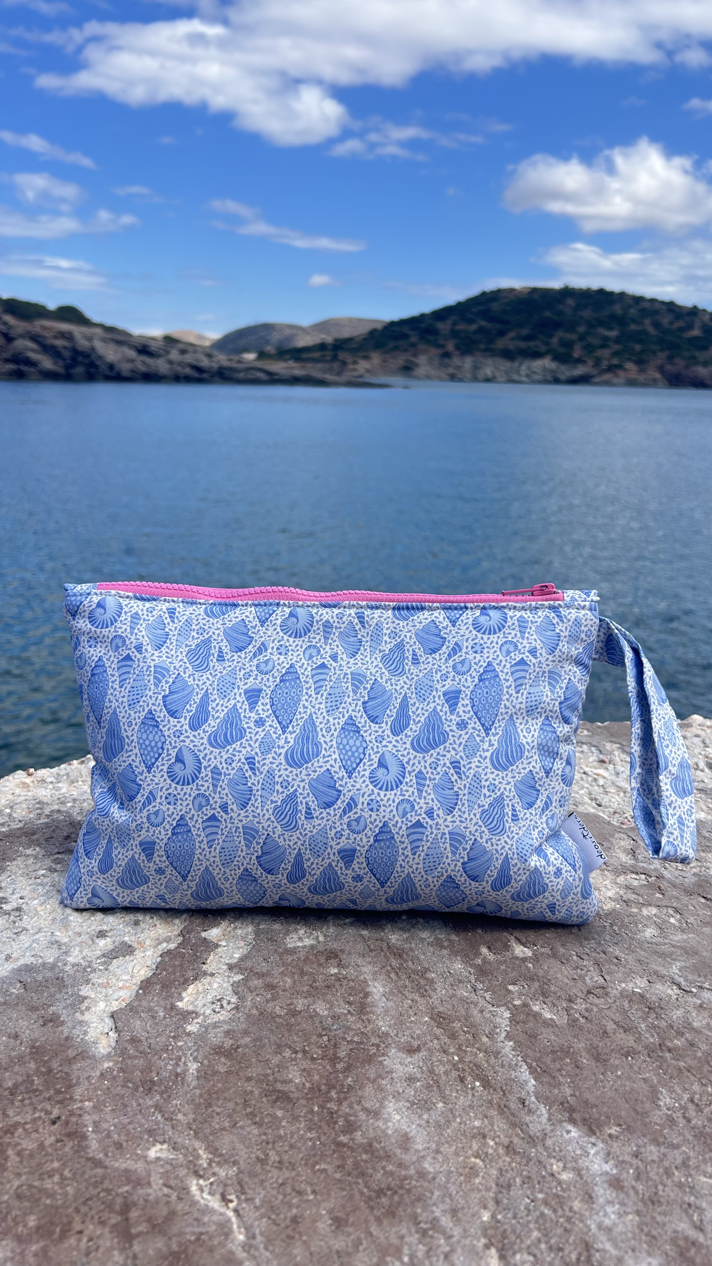 Blue and white seashell pattern pouch with pink zipper on a rock by the sea